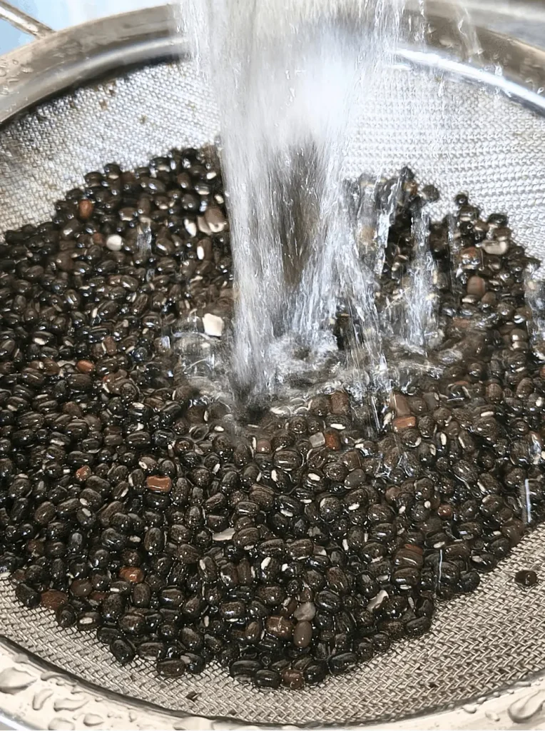Black lentils being rinsed under running water in a fine mesh strainer to remove dust and debris.