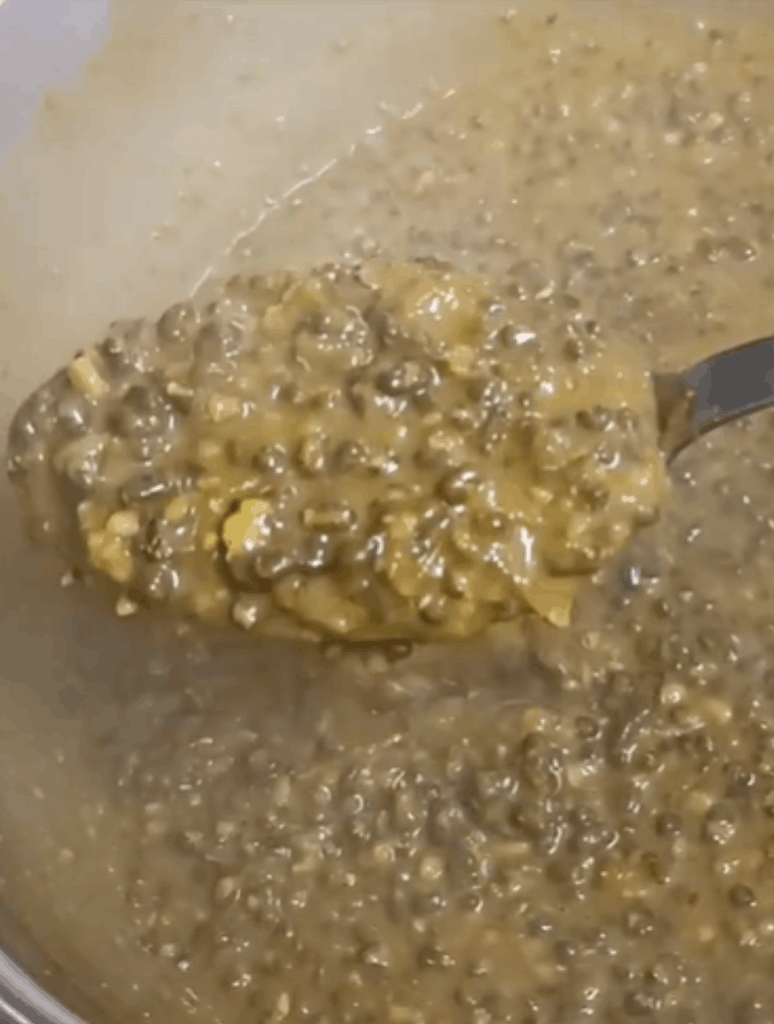 A spoonful of cooked black lentils lifted from a simmering pot, showing a thick, saucy texture.